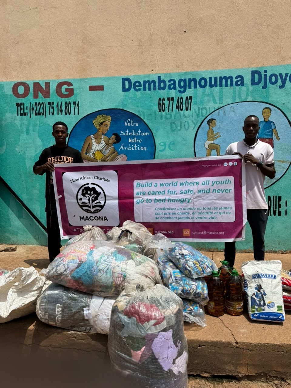Two volunteers hold the MACONA banner behind bags of clothing, bottles of cooking oil, pasta, and powdered milk