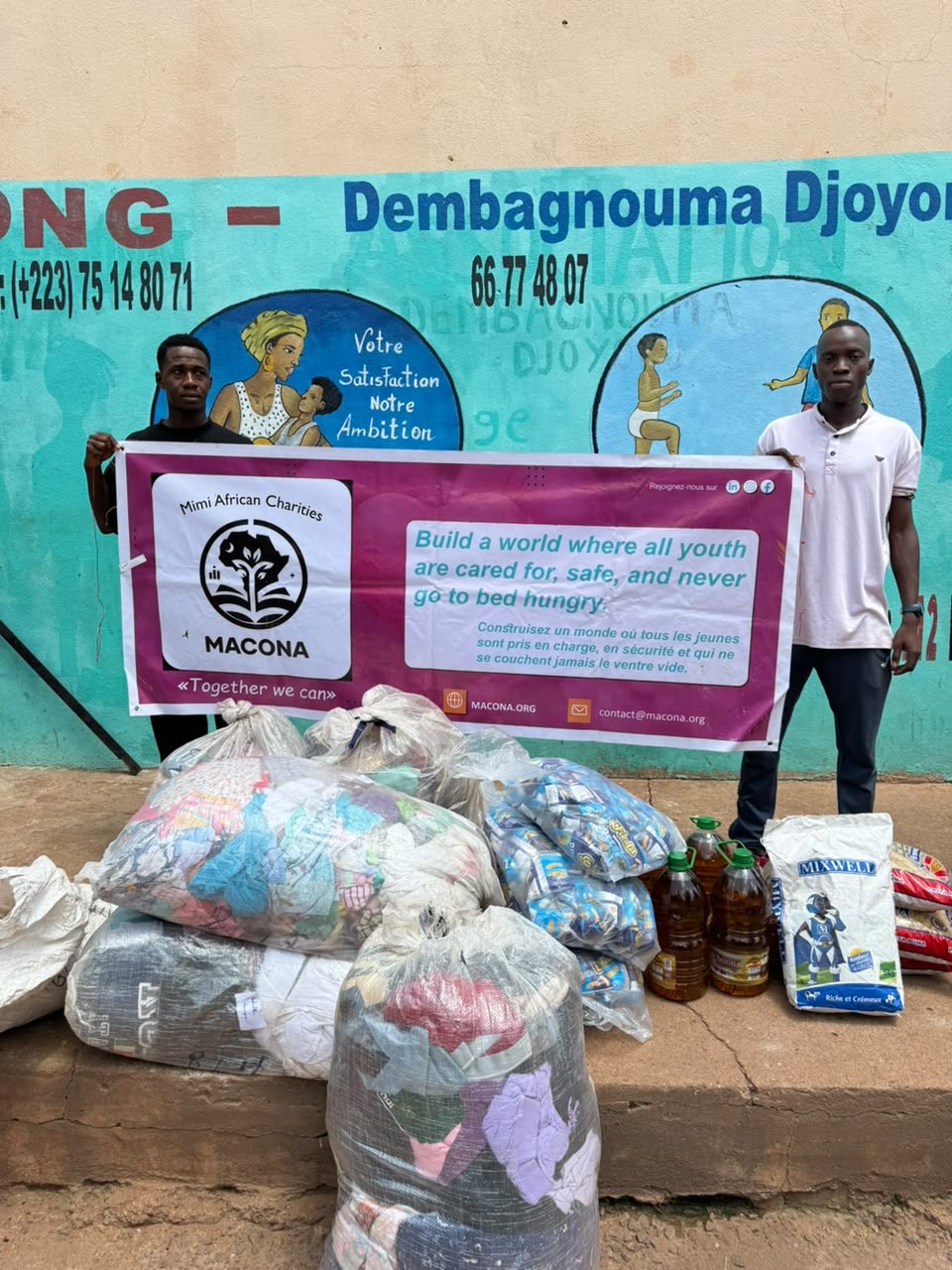 Two volunteers hold the MACONA banner behind bags of clothing, bottles of cooking oil, pasta, and powdered milk