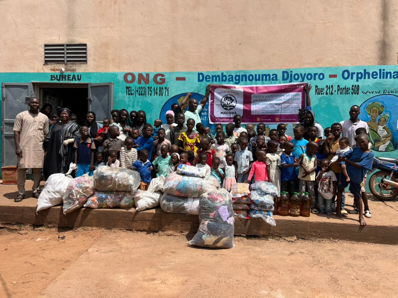 Children and staff of the ONG Dembagnouma Djoyoro Orphelinat gathered behind the delivered bags of clothing, food, and supplies in Bamako, Mali, with the MACONA banner held overhead