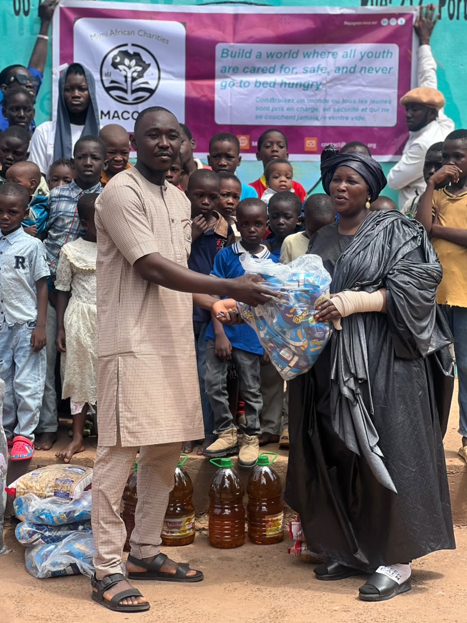 A MACONA volunteer hands a sealed bag of supplies to the director of the orphanage as the children look on