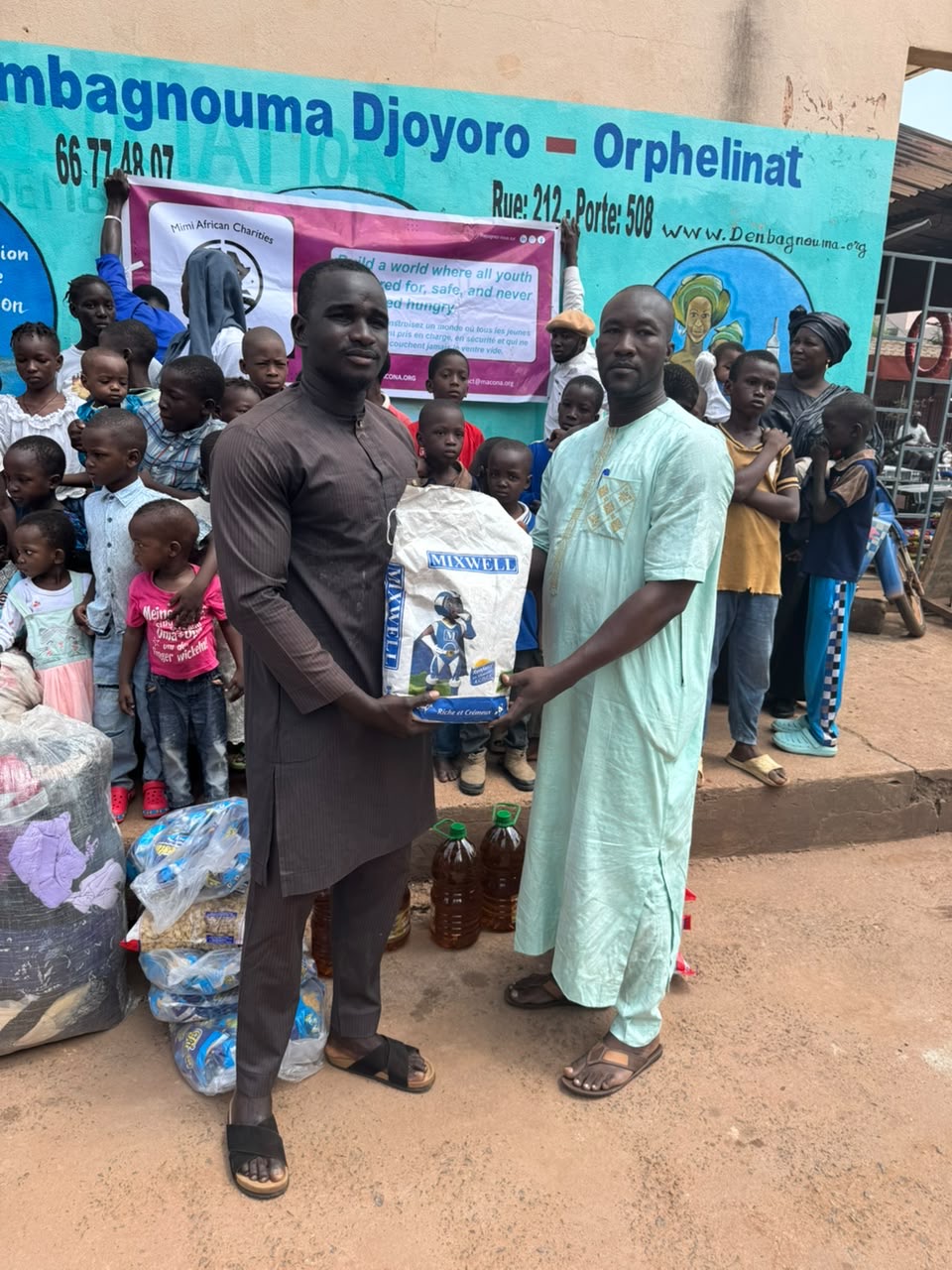 Two men hold a sack of powdered milk in front of the painted orphanage sign on Rue 212, Porte 508 in Bamako
